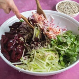 A bowl of salad with shredded beets, radishes, apples, greens, and sunflower seeds, being tossed with wooden utensils.