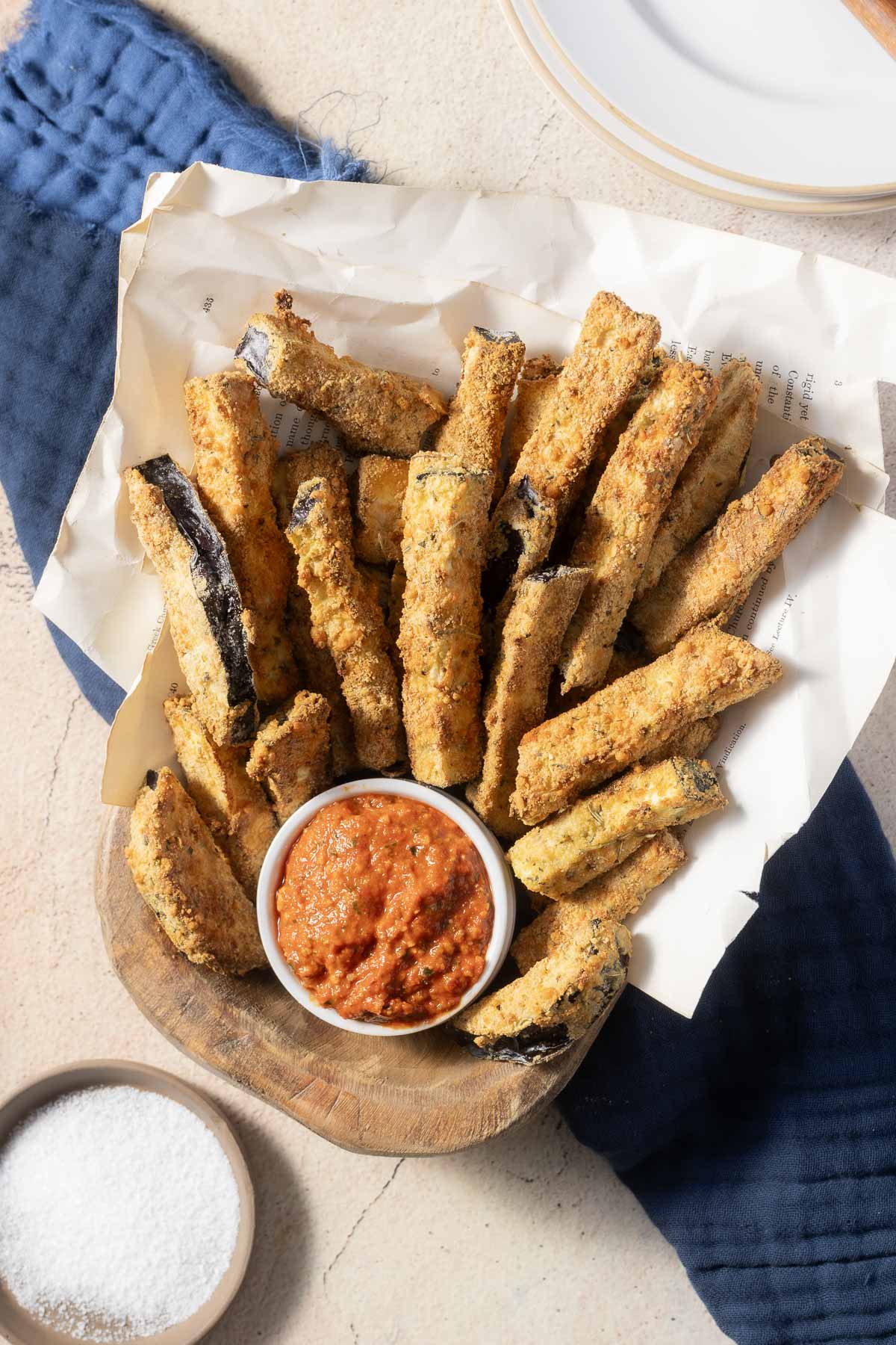 A basket of crispy eggplant fries with a small bowl of red dipping sauce on a wooden board, next to a bowl of salt and a blue cloth.