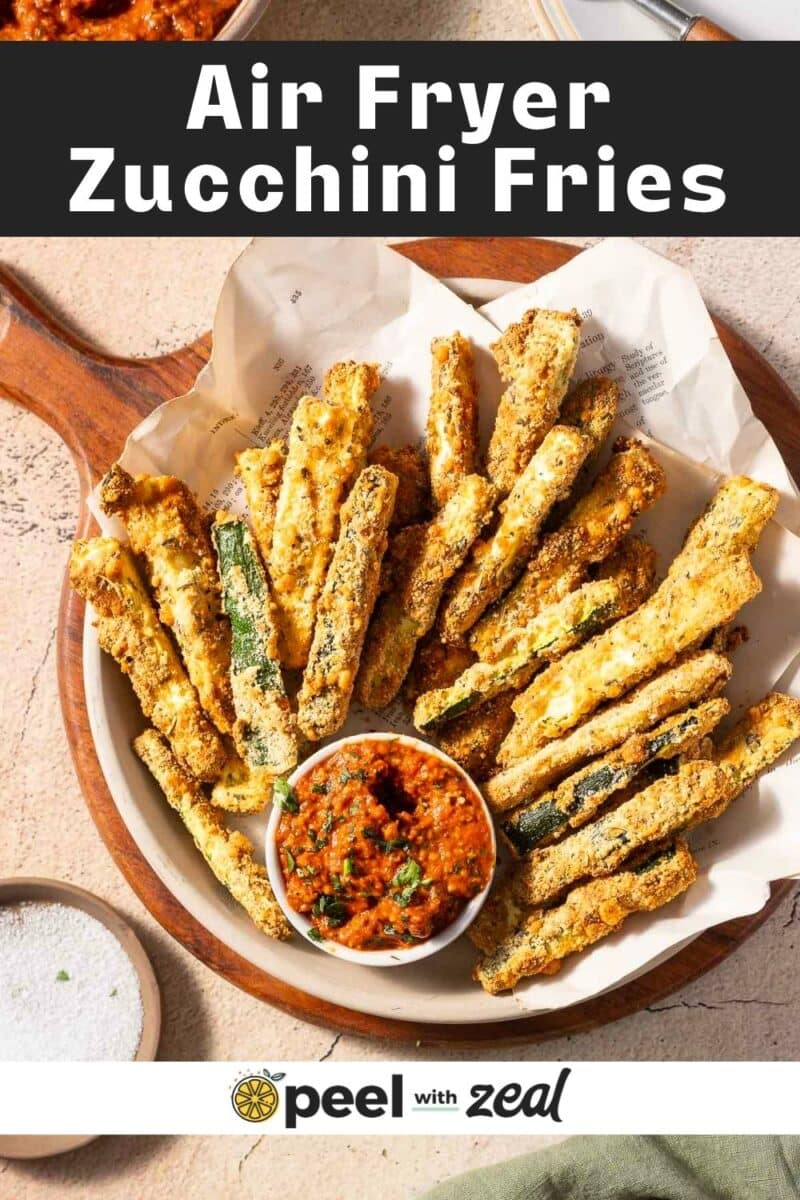 A bowl of air fryer zucchini fries with a small dish of red dipping sauce on a wooden serving tray. Text reads "Air Fryer Zucchini Fries.