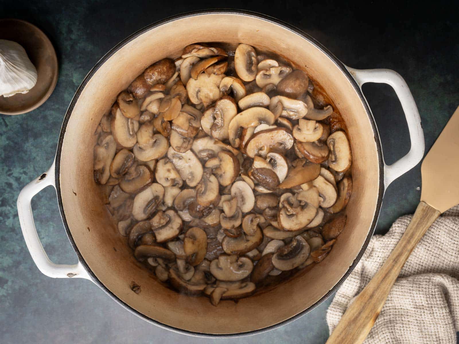 Sliced mushrooms cooking in a white pot on a dark countertop, surrounded by a wooden spatula, a small dish, and a cloth.