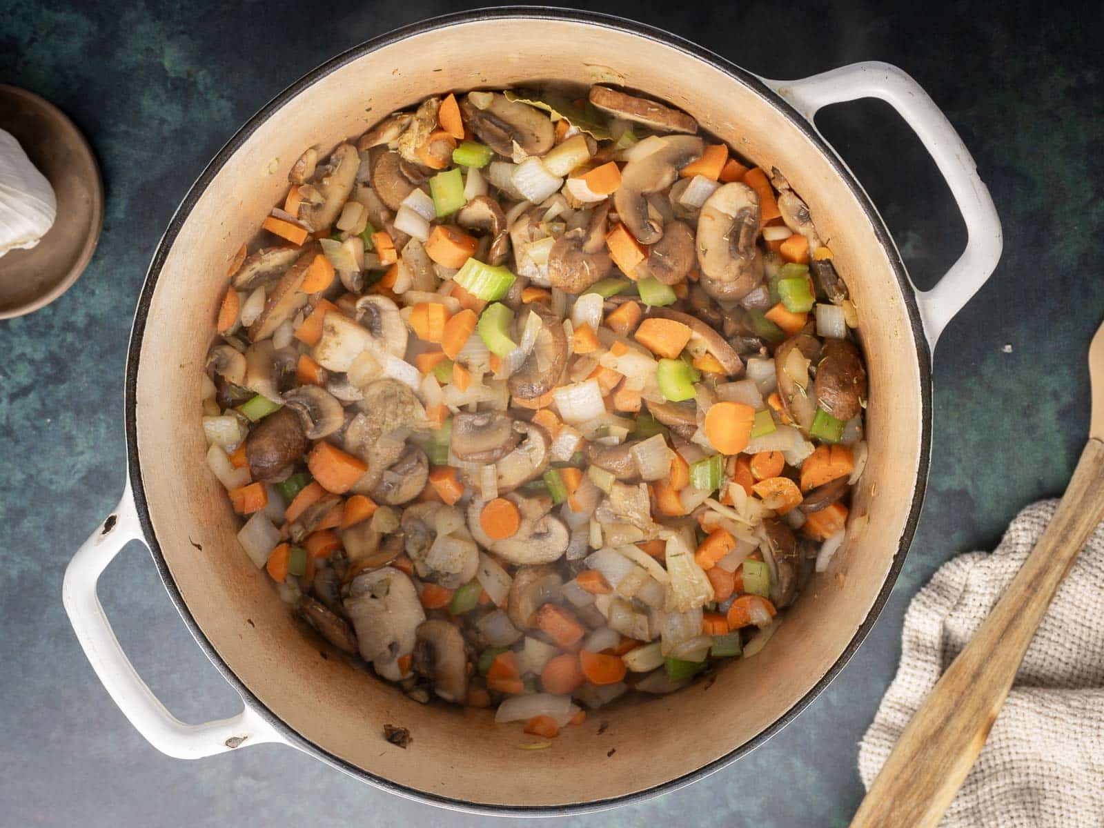 A pot filled with sautéed mushrooms, carrots, celery, and onions on a dark surface, with a wooden spoon and a cloth nearby.