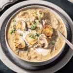 A bowl of creamy mushroom soup garnished with sliced mushrooms and chopped herbs, placed on a dark plate with a spoon on the side.