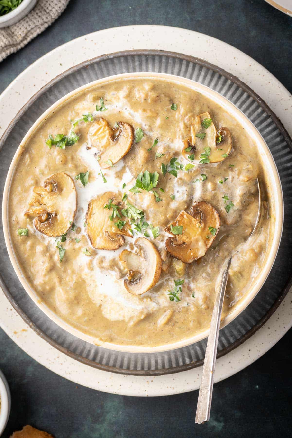A bowl of creamy mushroom lentil soup garnished with sliced mushrooms and fresh herbs, with a spoon placed inside.