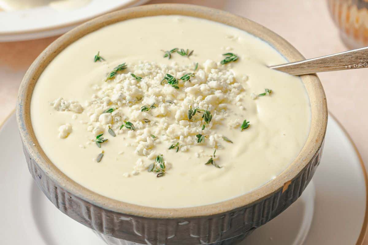 A bowl of creamy soup garnished with crumbled cheese and fresh herbs, with a spoon resting inside.
