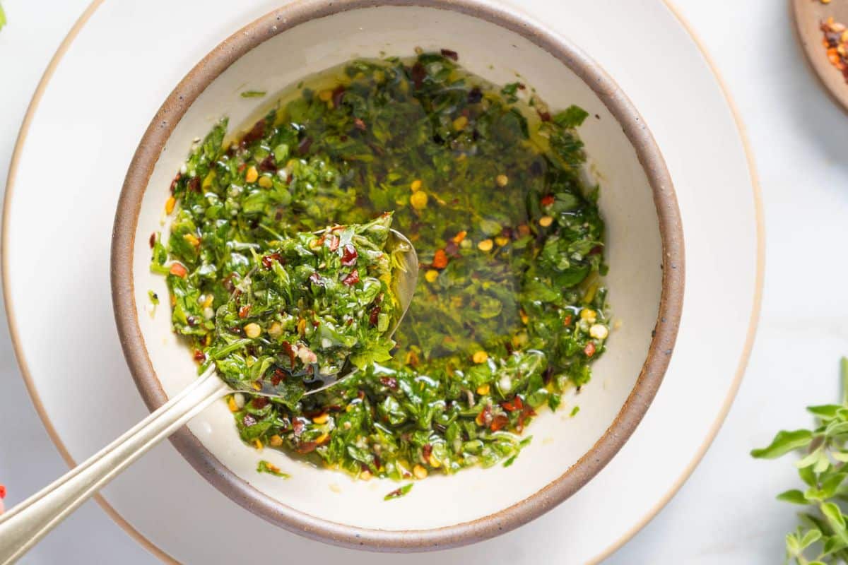 A bowl with green herb sauce, likely chimichurri, containing chopped herbs, oil, and red pepper flakes. A spoon rests in the sauce.