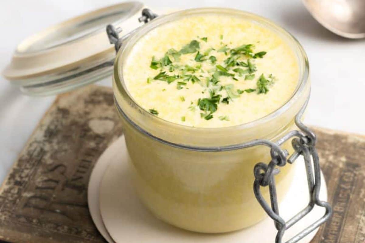 A jar filled with creamy garlic butter, garnished with chopped herbs, sits on a rustic book with a metal clasp.