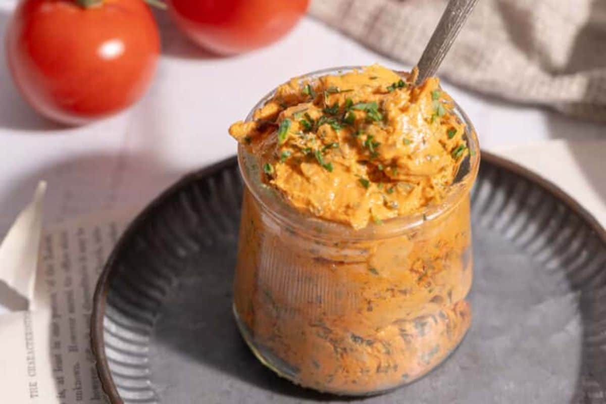 A jar filled with a creamy orange spread topped with herbs, placed on a round plate. In the background, there are tomatoes and a cloth.