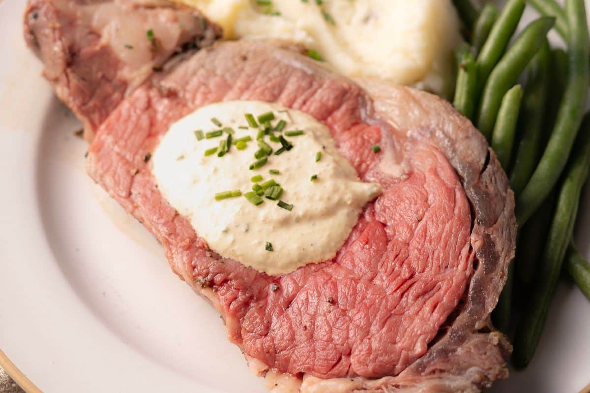 A plate with a slice of prime rib topped with creamy horseradish sauce and chives, alongside mashed potatoes and green beans.