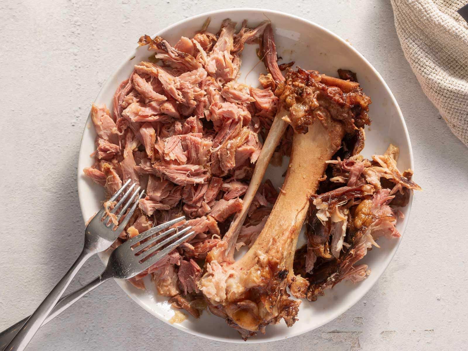A plate with shredded cooked meat and a large bone, accompanied by two metal forks on a light-colored surface.
