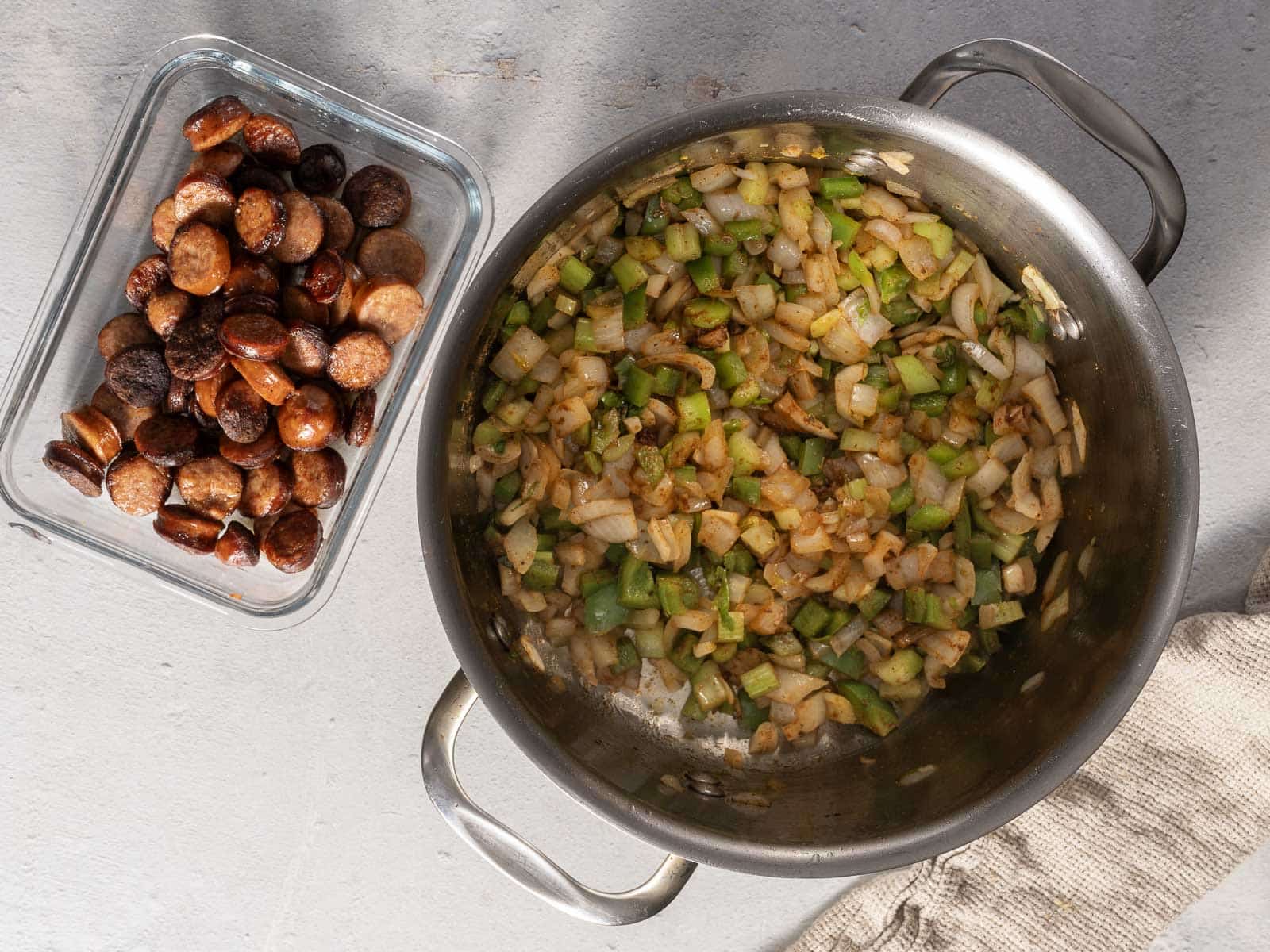 Chopped onions and celery sautéing in a pan next to a dish of cooked andouille sausage.