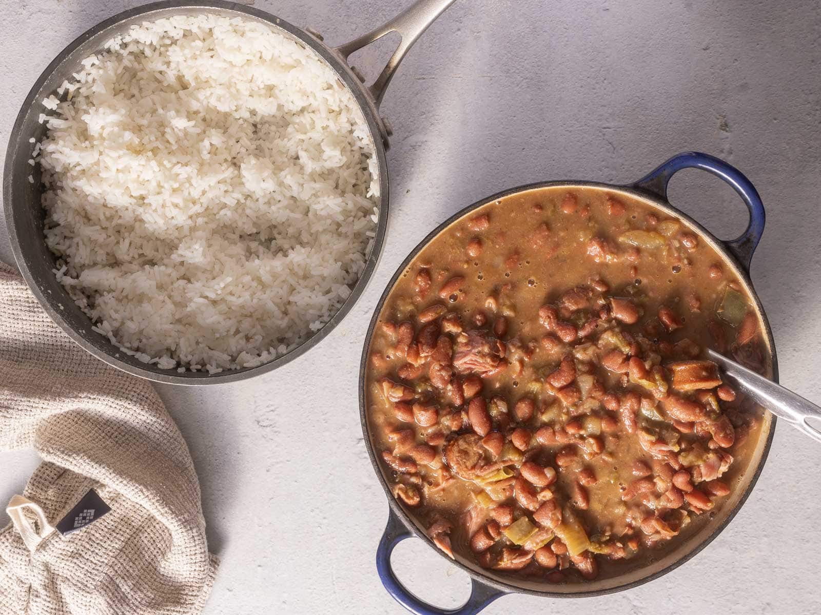A pot of cooked white rice next to a pot of red beans stew on a gray surface.