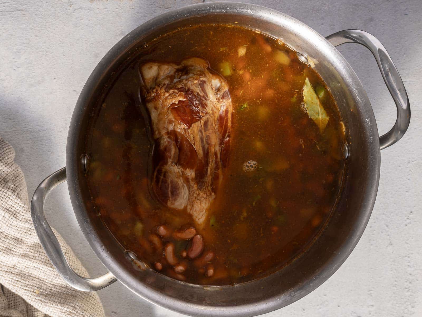 Pot of soup with a large meat piece, beans, and vegetables, simmering in broth.