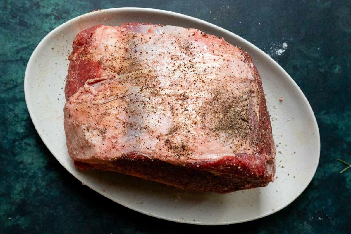 Raw seasoned beef roast on a white oval plate against a dark green background.
