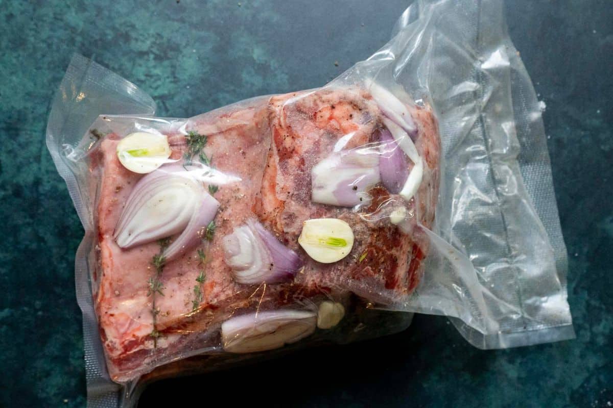 Vacuum-sealed package containing raw meat, sliced garlic, onion, and herbs on a dark green surface.