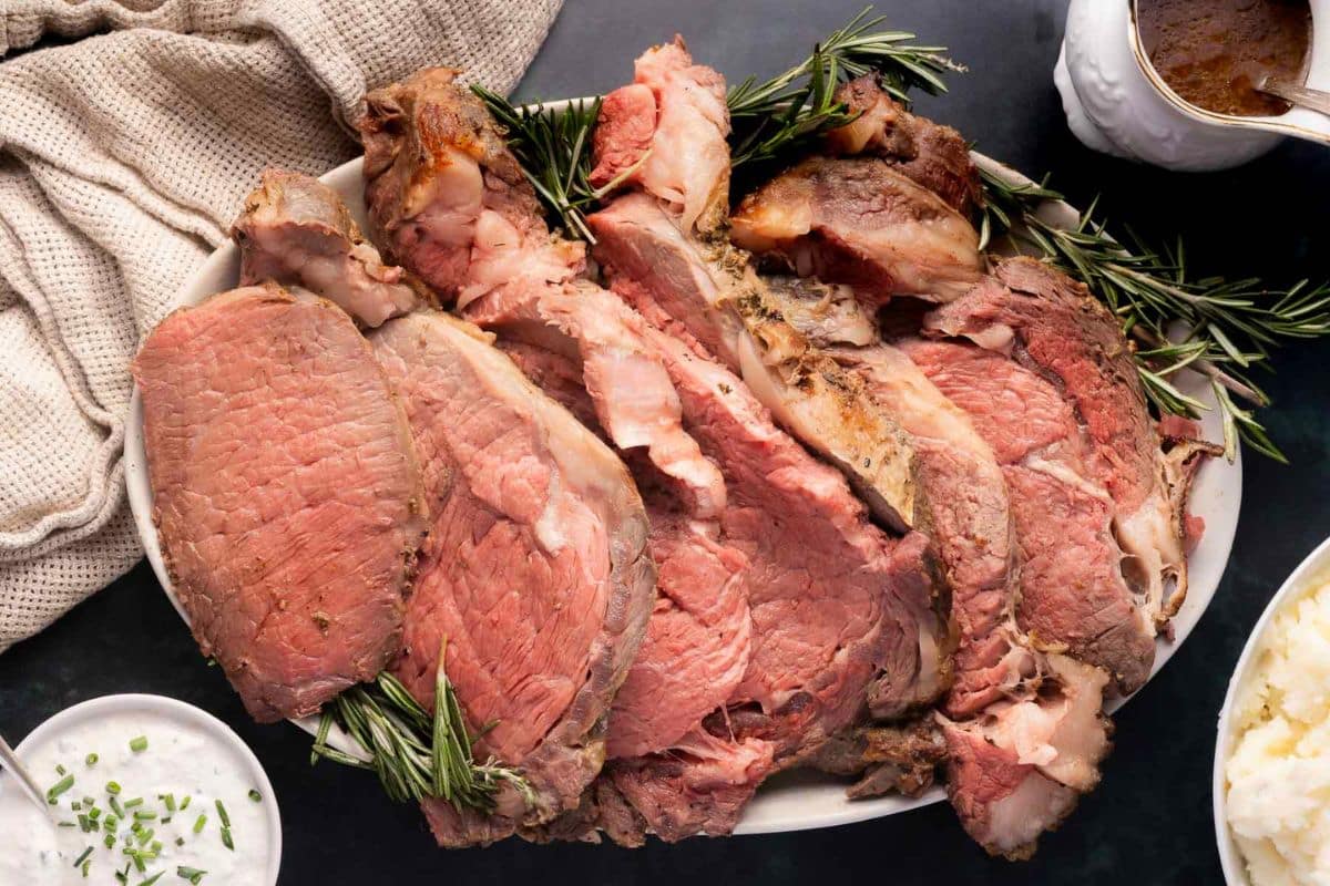 A platter of sliced roast beef garnished with rosemary, with a bowl of mashed potatoes and a napkin nearby.
