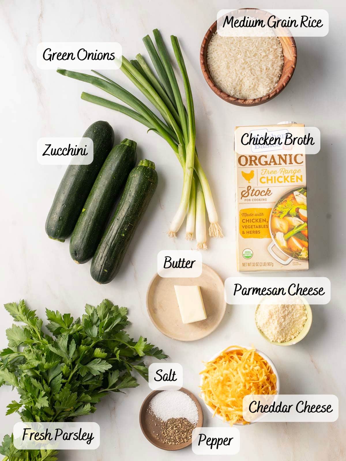 Ingredients on a counter including green onions, zucchini, medium grain rice, chicken broth, butter, Parmesan and cheddar cheese, salt, pepper, and fresh parsley.