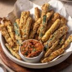 A bowl of crispy zucchini fries with a small dish of red dipping sauce garnished with herbs.