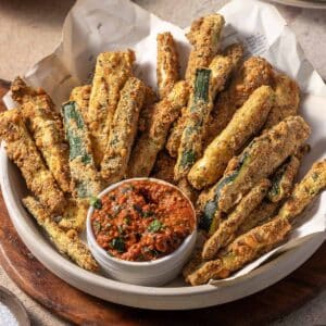 A bowl of crispy zucchini fries with a small dish of red dipping sauce garnished with herbs.