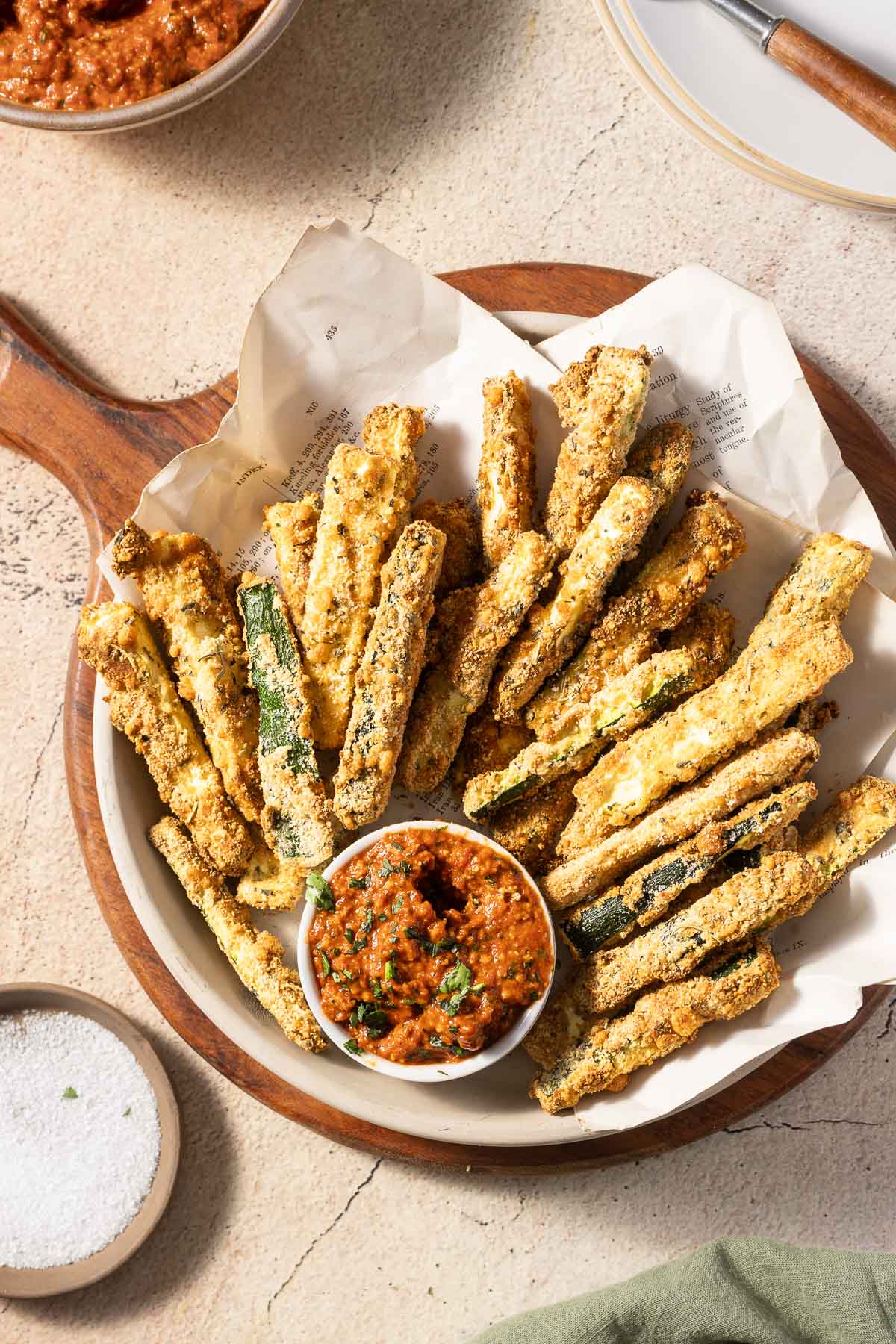 A plate of crispy zucchini fries is arranged on parchment, accompanied by a small bowl of red dipping sauce. A small bowl of coarse salt is nearby on a wooden surface.