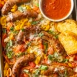 Baked Italian sausage and peppers on a tray with onions, topped with cheese and basil delight the senses. Accompanied by a bowl of rich red sauce and slices of garlic bread, this dish is a flavorful feast.