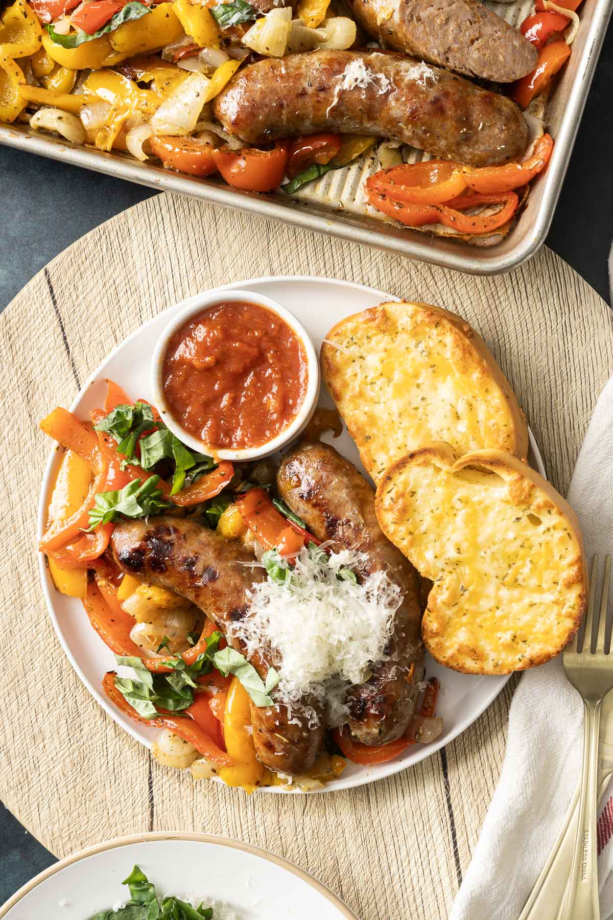 A plate of baked Italian sausages with roasted bell peppers and onions, topped with grated cheese, sits next to a small dish of sauce and toasted bread slices.