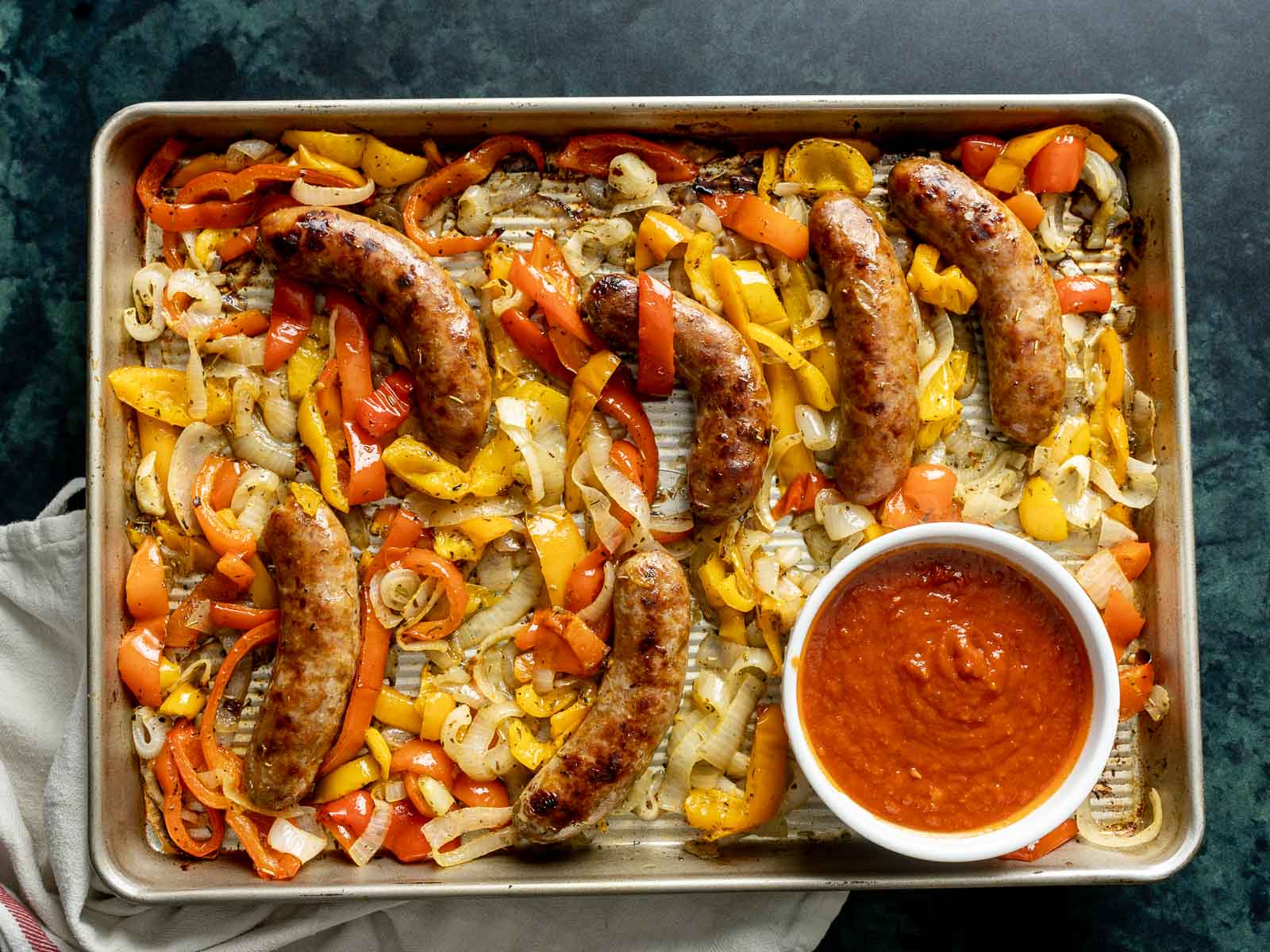 A baking tray features a delicious spread of baked Italian sausage alongside sliced bell peppers and onions, perfectly roasted. A bowl of rich red sauce sits invitingly on the side.