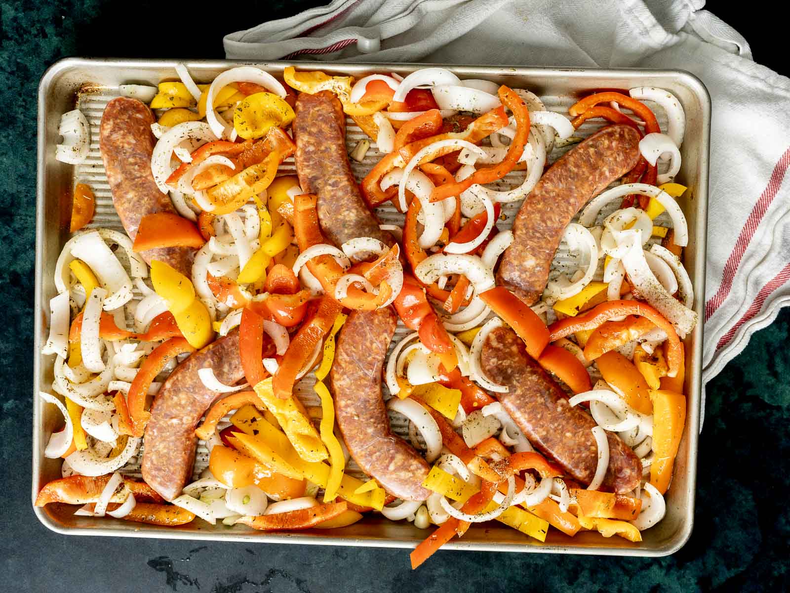 Sliced onions and bell peppers with baked Italian sausages are arranged on a baking tray, ready for cooking, on a dark countertop with a cloth beside.