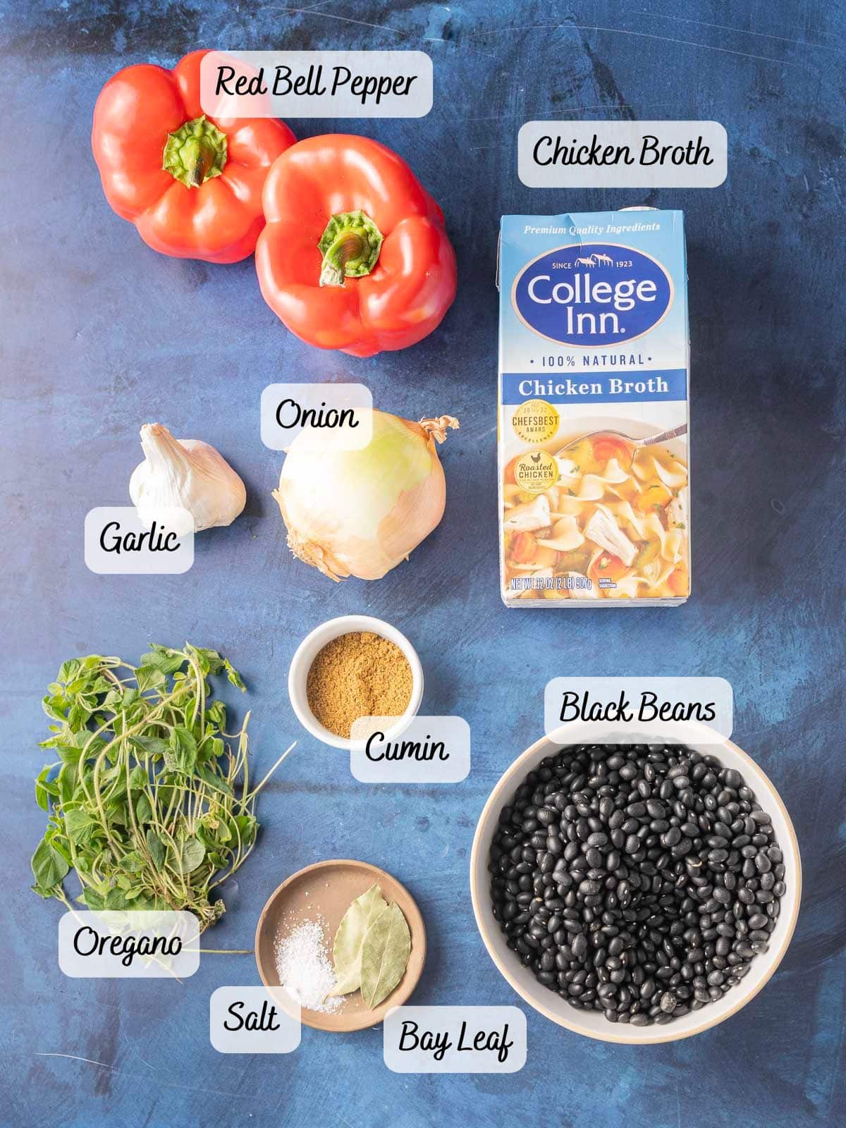 Ingredients arranged on a blue surface include red bell pepper, chicken broth, onion, garlic, black beans, cumin, oregano, salt, and a bay leaf.