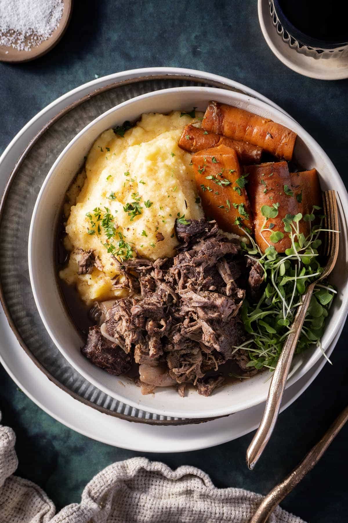 A bowl containing mashed potatoes, shredded beef, and roasted carrots, garnished with fresh herbs, on a dark surface. A fork rests inside the bowl.