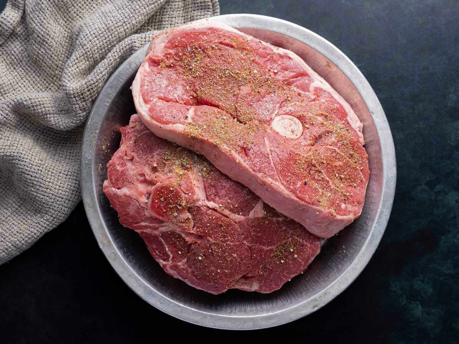 Raw seasoned pork steaks in a metal bowl, next to a gray cloth on a dark surface.