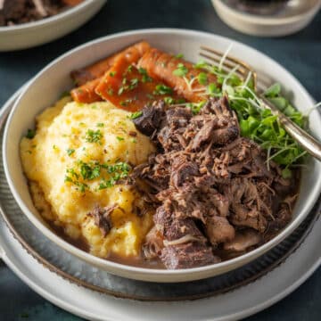 A bowl with polenta, tender braised lamb, sliced carrots, and greens, garnished with herbs and a hint of red wine, on a plate with a fork.