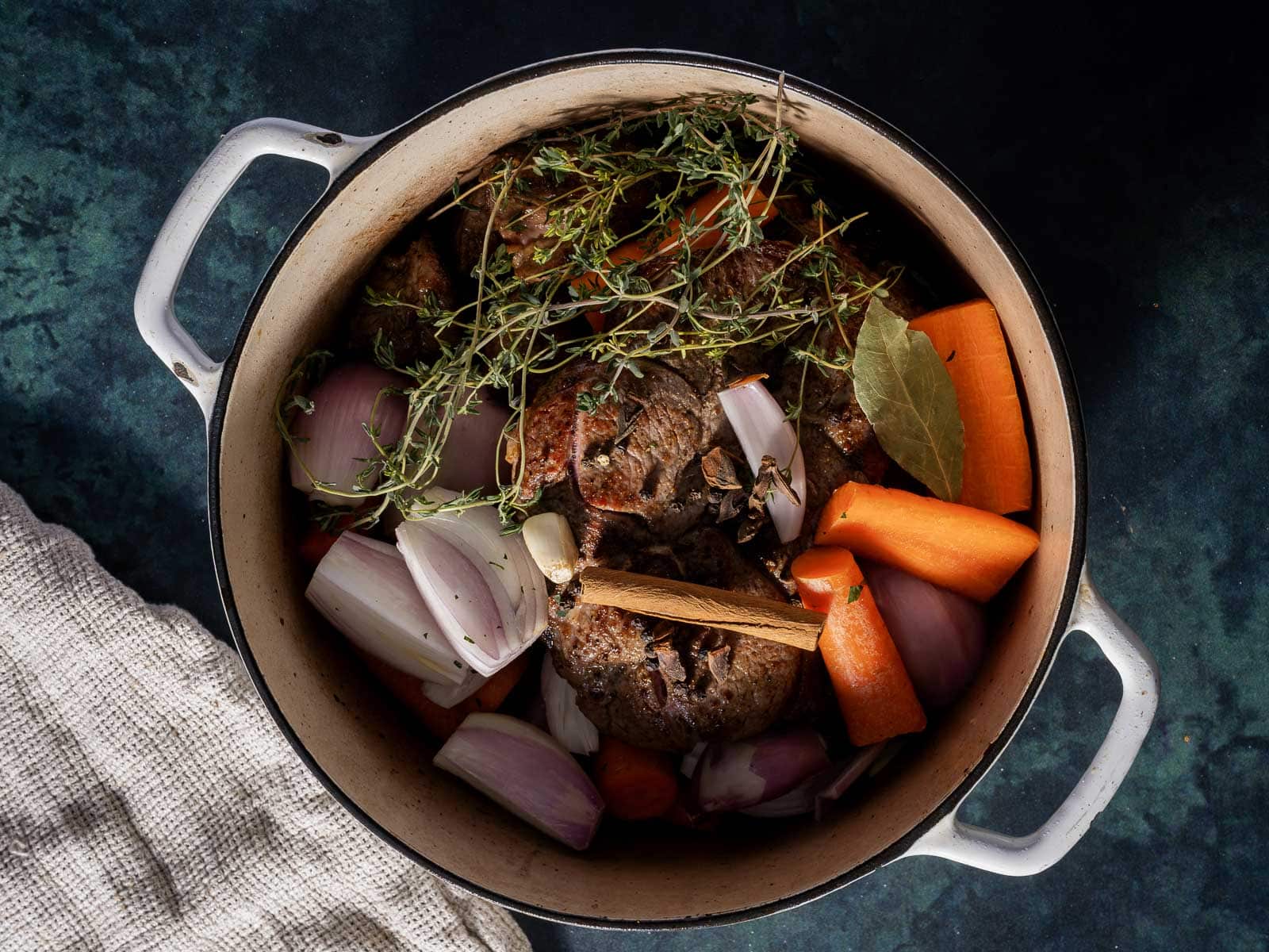 A pot filled with seasoned beef chunks, carrots, onions, garlic, thyme, and a bay leaf, ready for cooking.