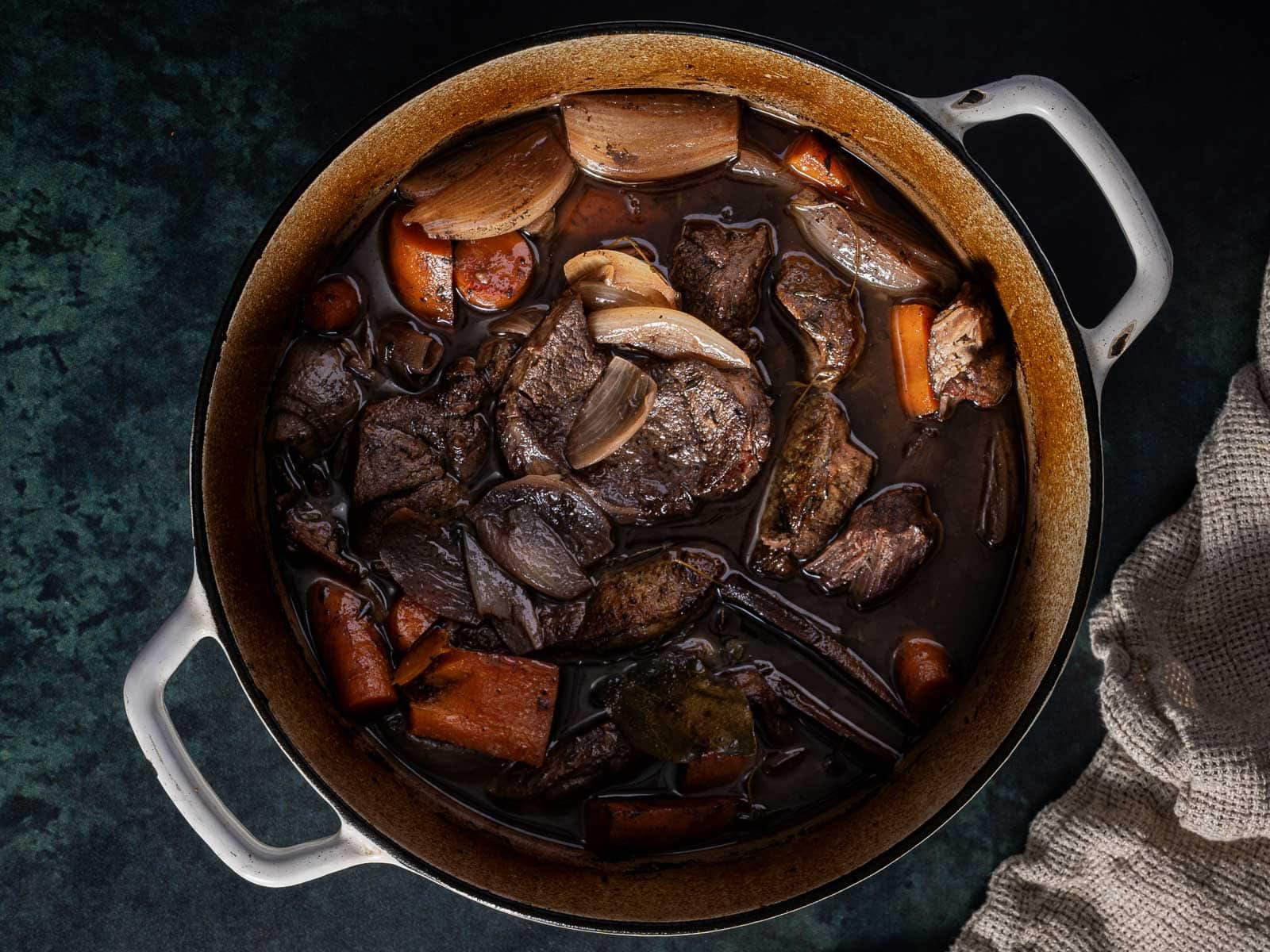 A pot of beef stew with chunks of beef, carrots, and onions in a dark broth on a textured dark surface.