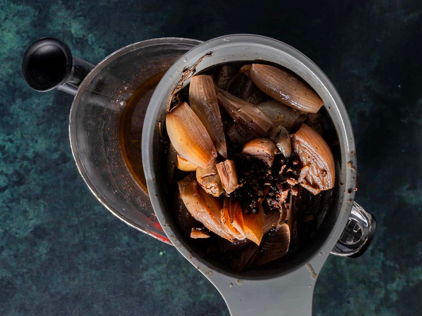 A strainer containing cooked onion and spices rests over a glass measuring cup filled with dark liquid on a dark surface.
