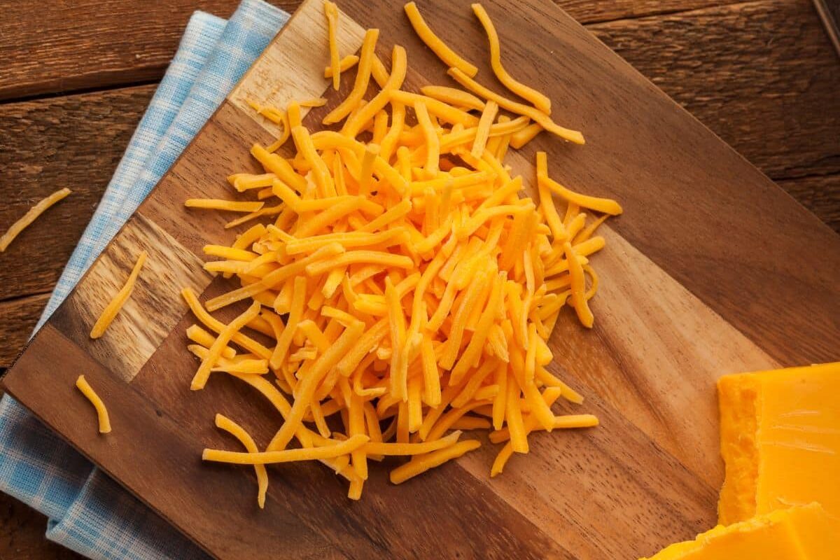 A pile of shredded cheddar cheese, perfect for your bean and cheese tacos, rests on a wooden cutting board with a blue and white cloth underneath.