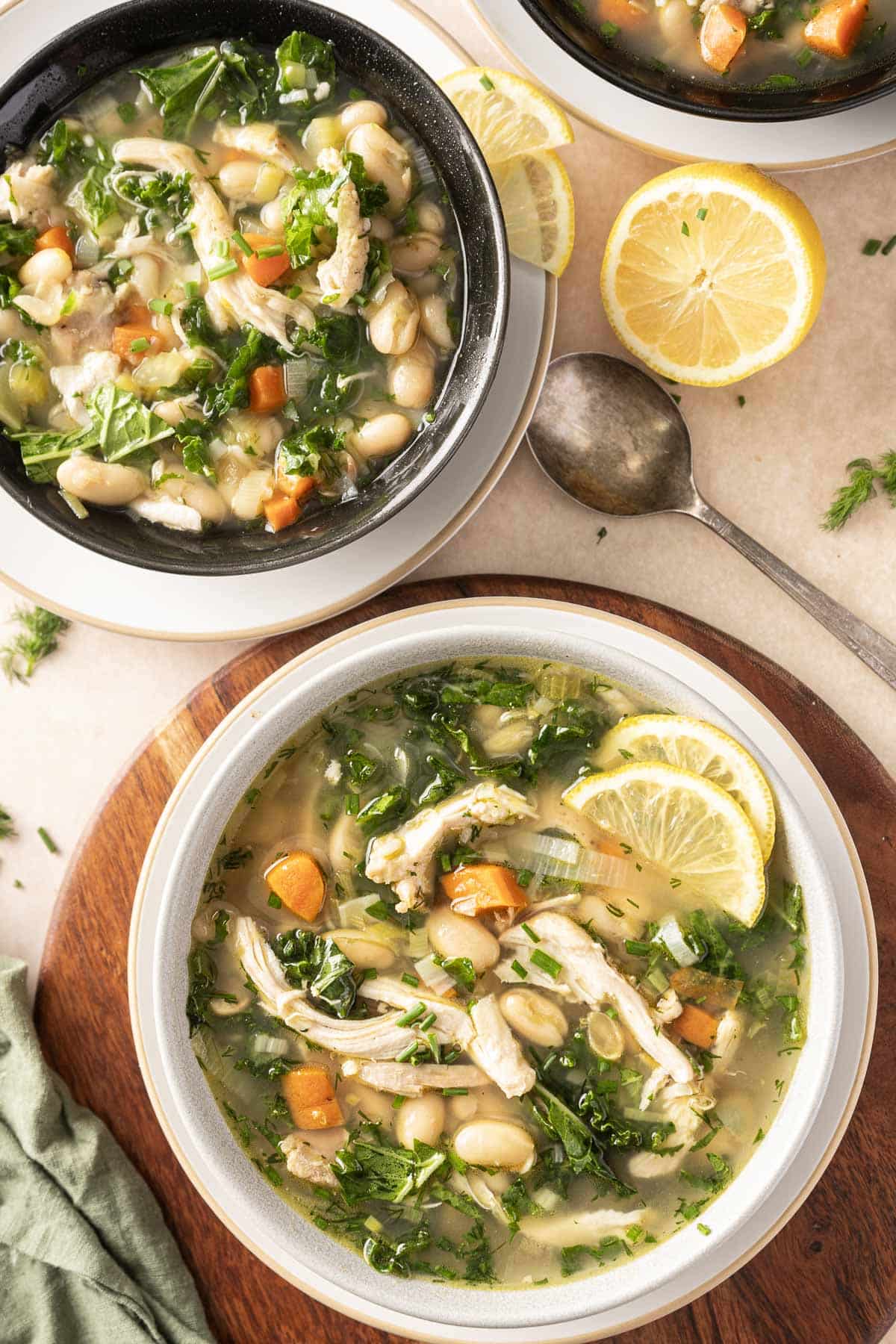 Two bowls of hearty chicken and white bean soup with vibrant greens sit invitingly on the table, garnished with lemon slices. A spoon rests nearby, accompanied by fresh lemon wedges, ready to enhance the zesty flavors.