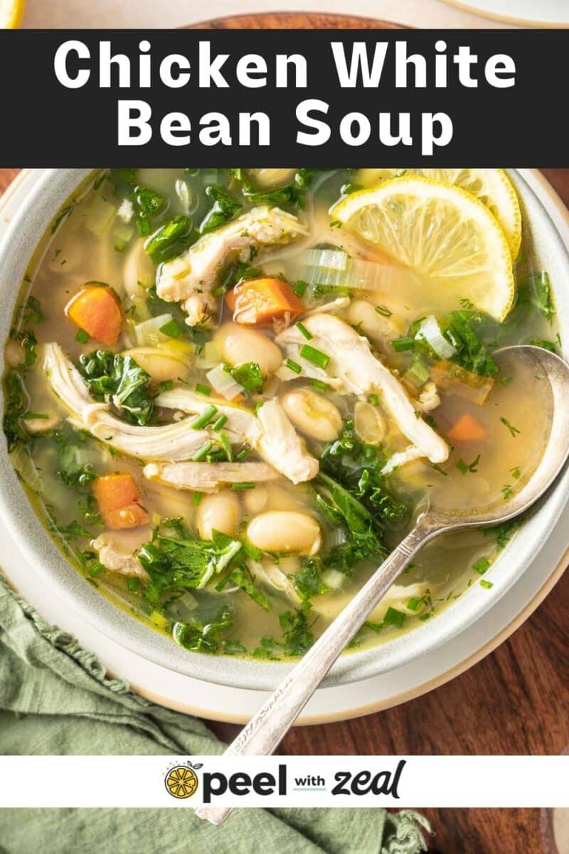A bowl of chicken white bean soup with carrots, greens, and a lemon slice, accompanied by a spoon.