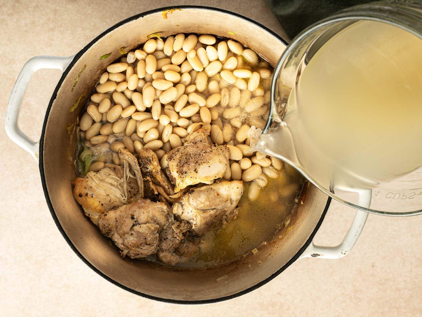 A pot with cooked chicken pieces and white beans awaits as broth is poured in from a clear pitcher, creating a hearty chicken soup.