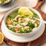 A bowl of chicken and white bean soup with vegetables and herbs, garnished with lemon slices, on a wooden board. Kale adds a vibrant touch, while a spoon rests invitingly in the bowl.