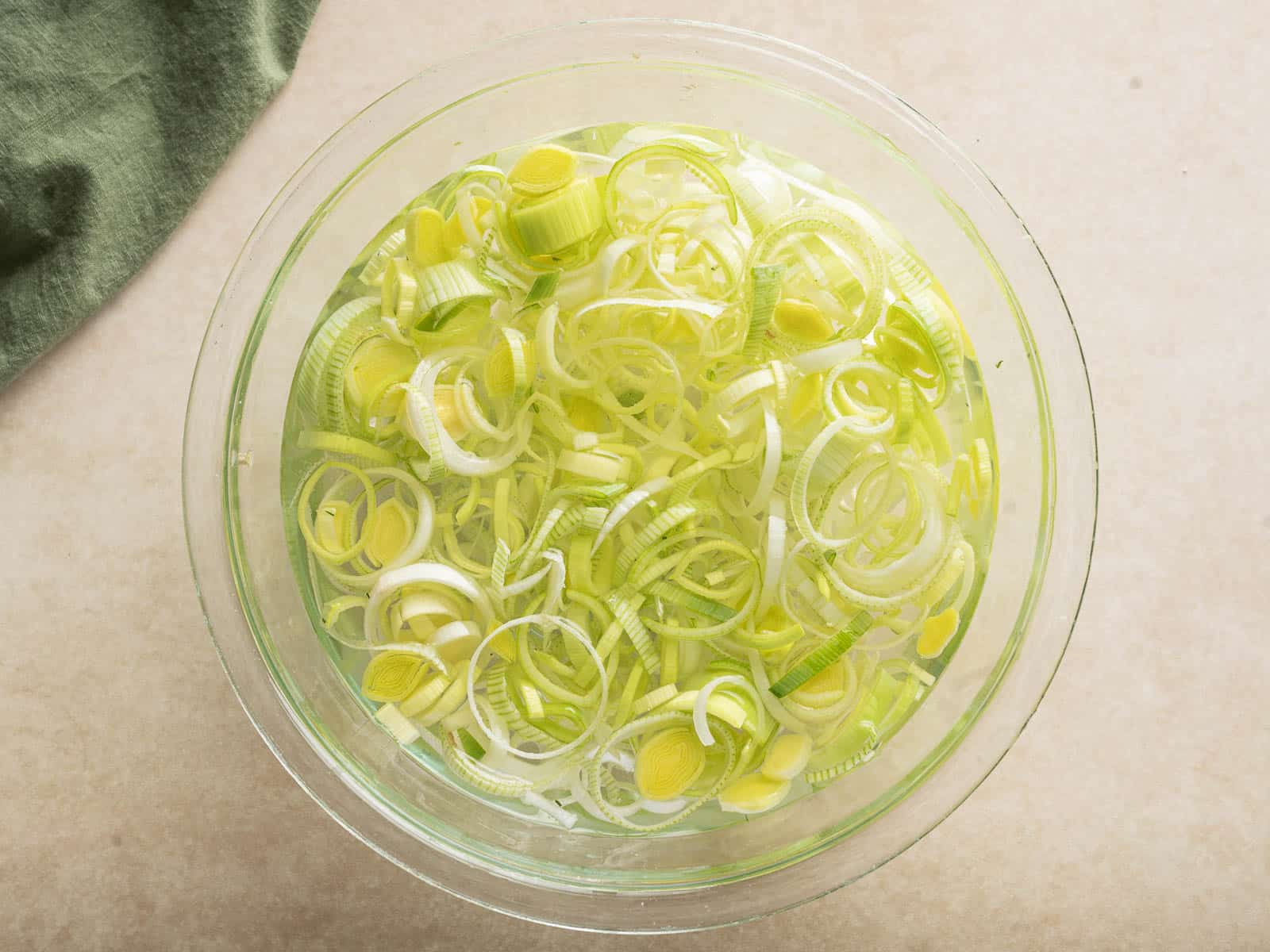 A bowl of sliced leeks soaking in water on a beige surface, with a green cloth nearby—a perfect start for your hearty white bean soup.
