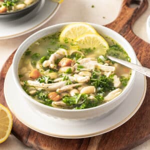 A bowl of chicken and white bean soup with vegetables and herbs, garnished with lemon slices, on a wooden board. Kale adds a vibrant touch, while a spoon rests invitingly in the bowl.