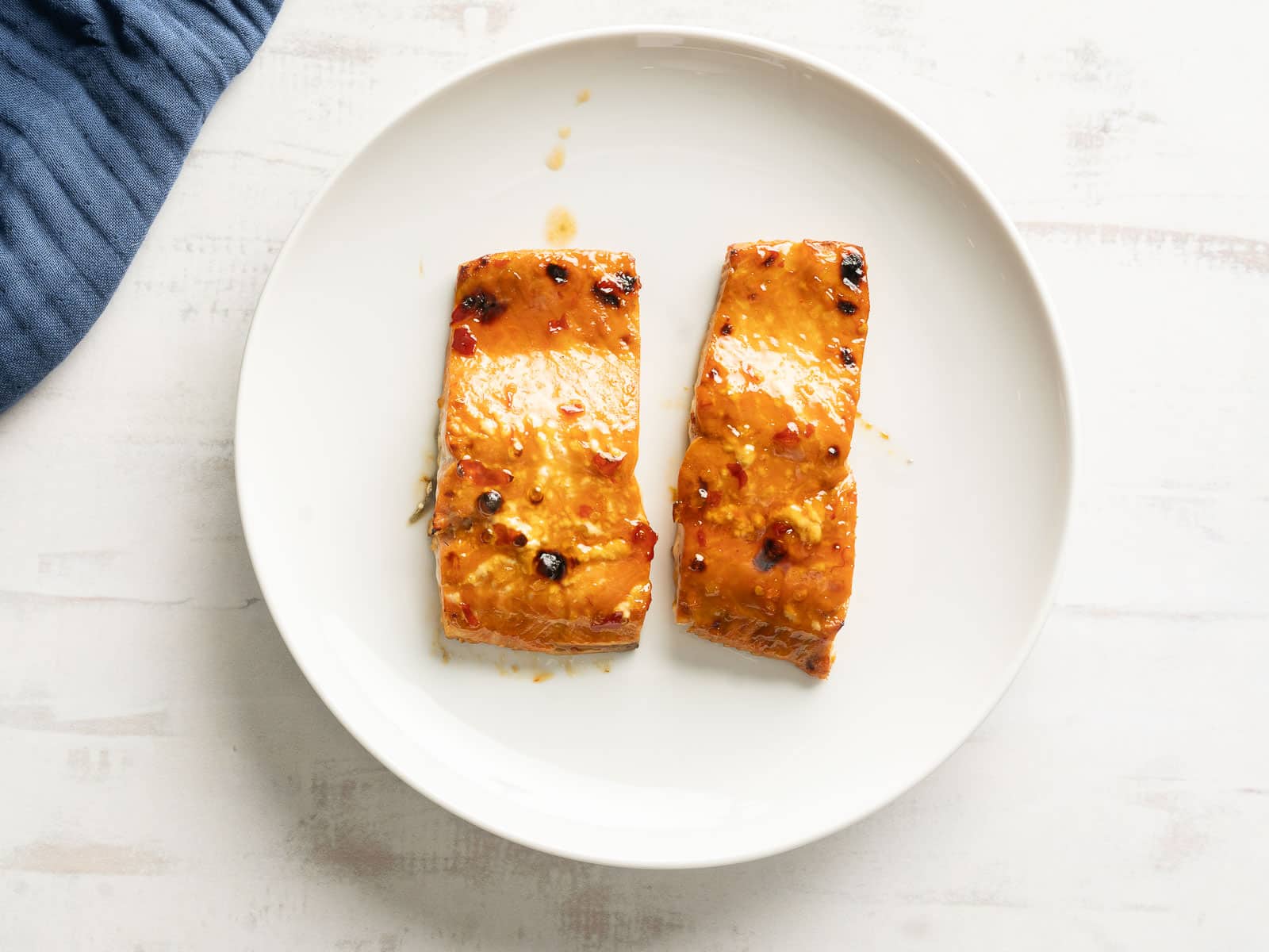 Two pieces of sweet chili salmon glisten on a white plate, accompanied by the soft hues of a blue cloth peeking from the side.