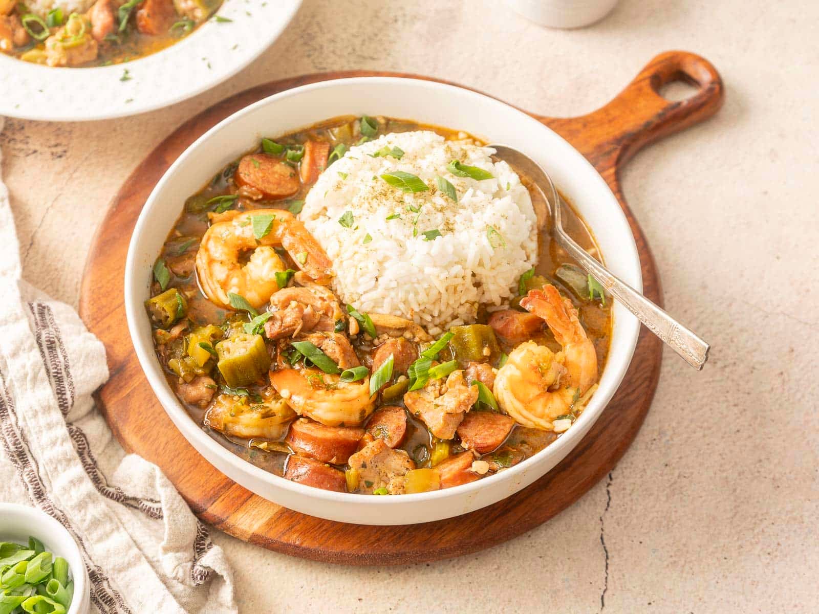 A bowl of gluten-free gumbo with shrimp, sausage, and vegetables, topped with a mound of white rice, sits on a wooden board.