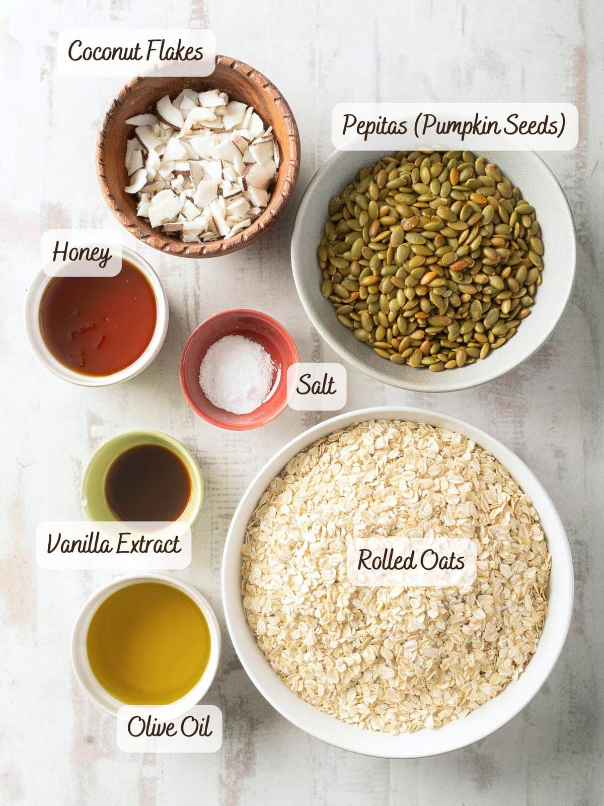 Various ingredients in bowls including coconut flakes, pepitas, honey, salt, vanilla extract, rolled oats, and olive oil, labeled and arranged on a light background.