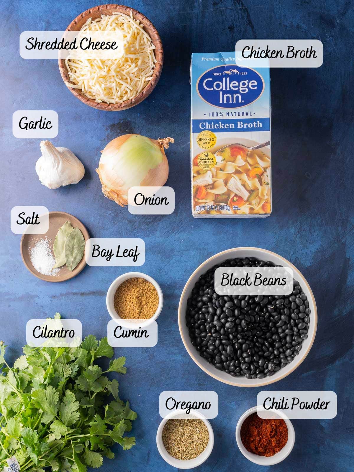 The ingredients for the recipe are laid out: a mix of shredded cheese, chicken broth, garlic, onion, salt, bay leaf, cilantro, cumin, black beans transforming into refried goodness with oregano and chili powder. 