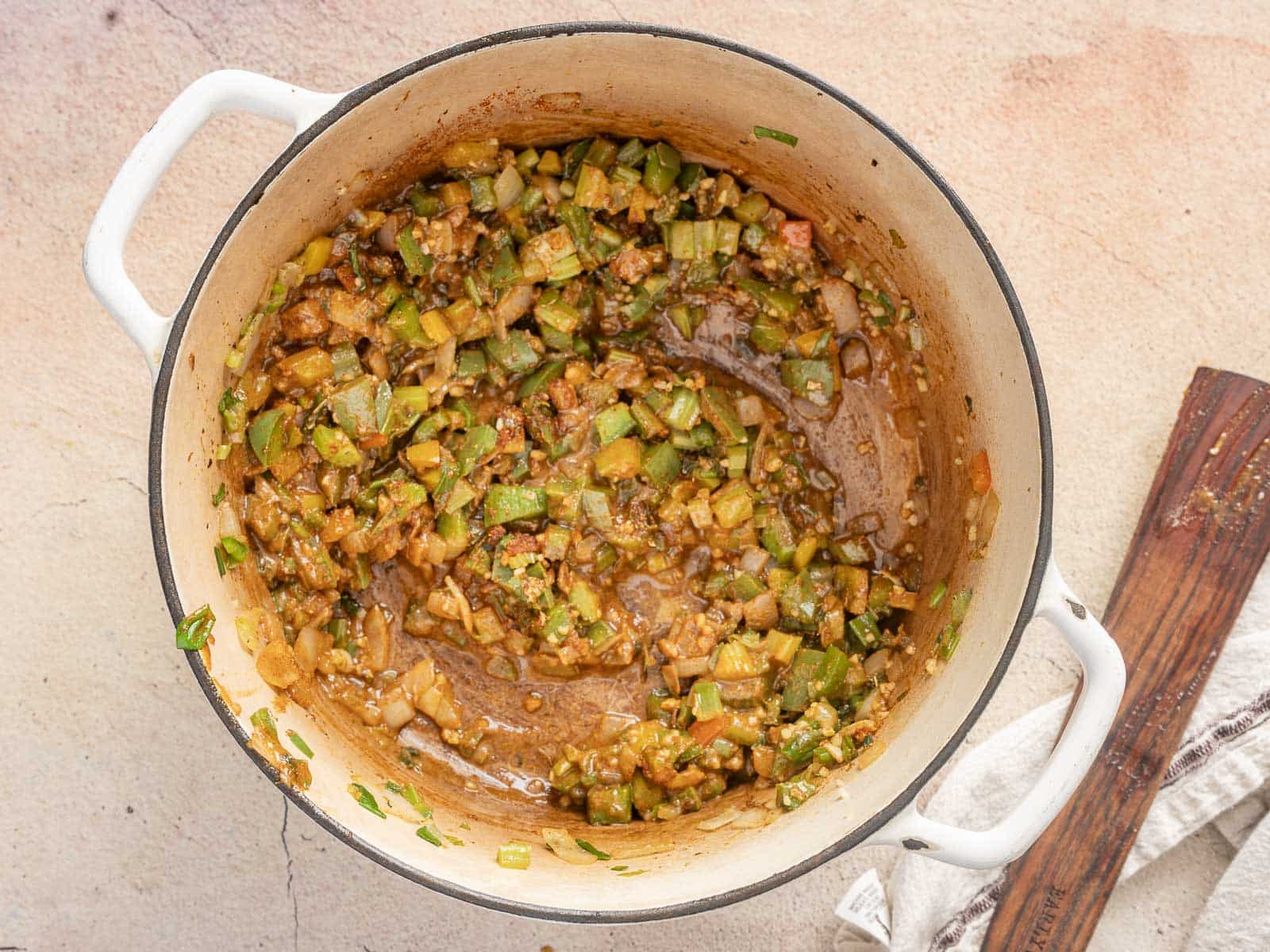 A white pot containing a gluten-free mixture of saut&eacute;ed vegetables, including green bell peppers and onions, sits on a countertop next to a wooden spatula and a cloth, reminiscent of the vibrant flavors found in a classic gumbo.