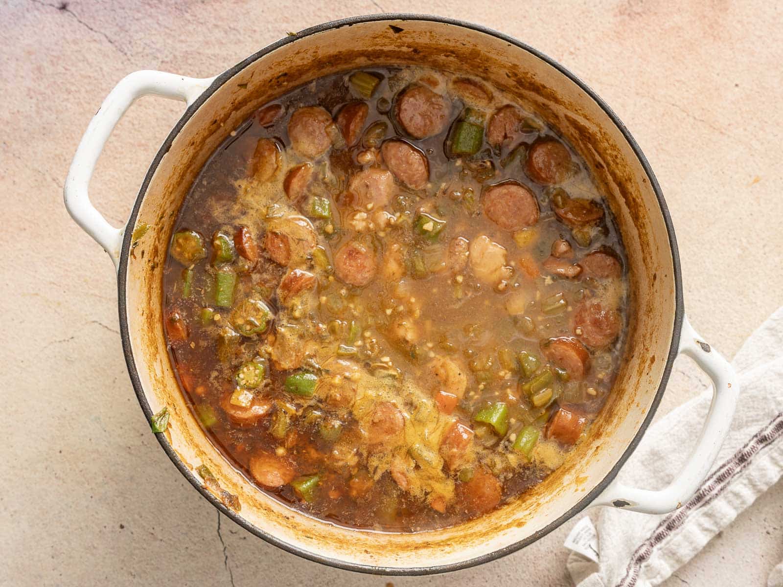 A pot of gluten-free gumbo with sliced sausage, okra, and a rich broth. The dish appears seasoned and ready to serve, with a white towel partially visible beside the pot.