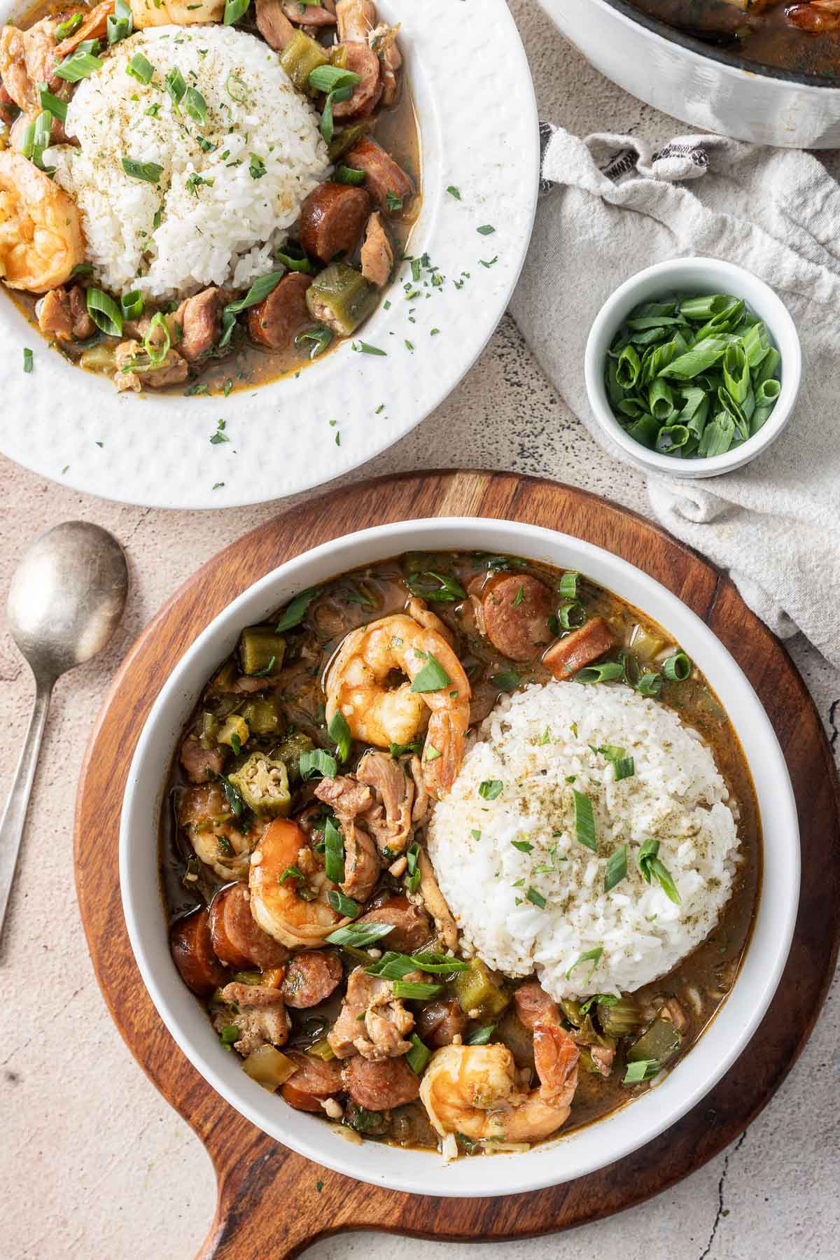 Gluten-free bowls of gumbo brimming with shrimp, sausage, and rice, garnished with green onions, are artfully arranged on a wooden board.