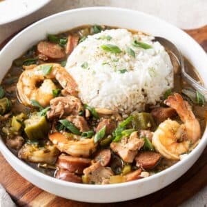 A bowl of gluten-free gumbo brimming with shrimp, sausage, okra, and chicken is served alongside a mound of white rice and garnished with vibrant green onions.