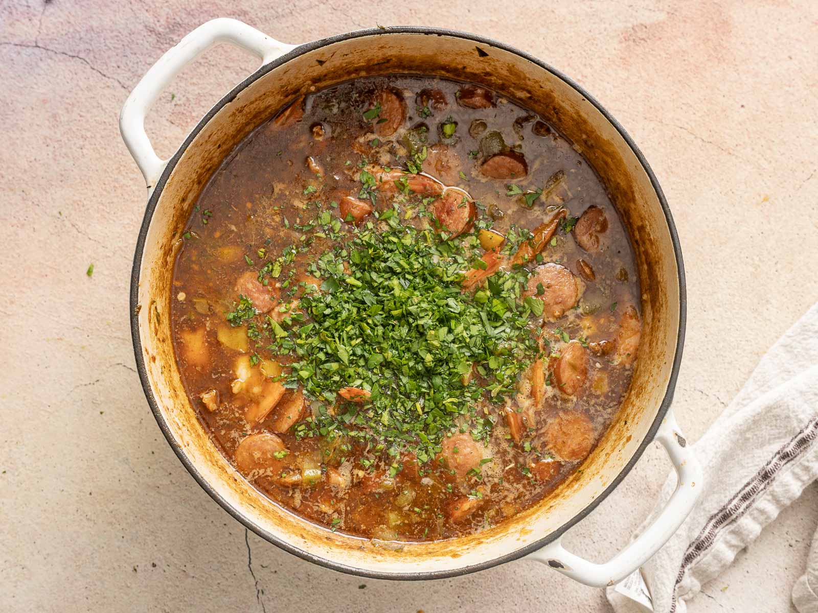 A pot of gluten-free sausage and vegetable gumbo, garnished with chopped herbs, is placed on a light-textured surface with a cloth nearby.